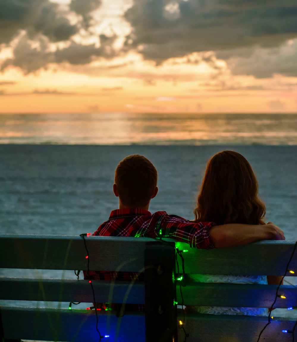 Couple watches the sunset from a bench wrapped in christmas lights