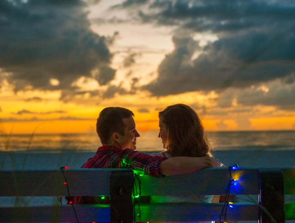 Couple at the beach for christmas photo with christmas lights wrapped around a bench